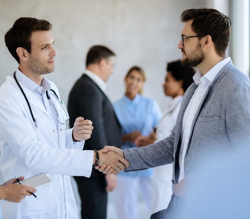 Male doctor handshaking with a businessman while meeting in a hallway at medical clinic.