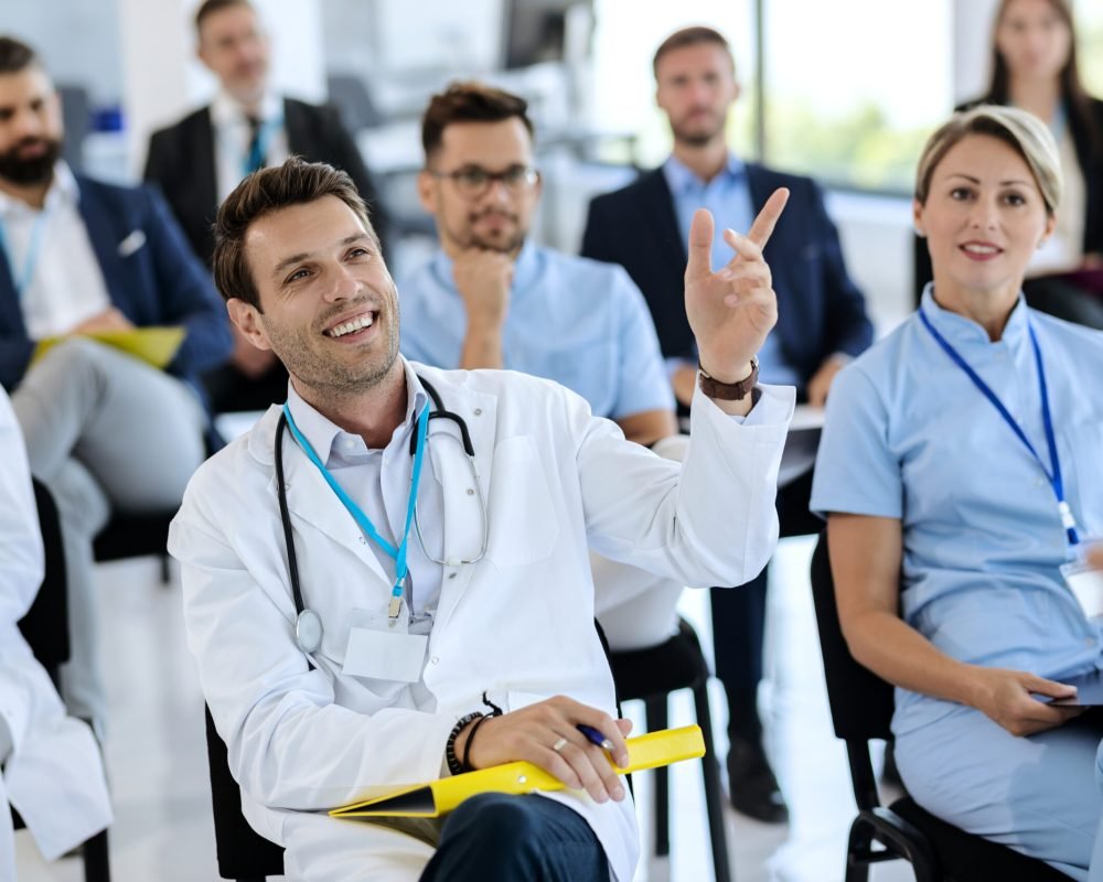Happy doctor attending a seminar and raising his hand to ask a question at convention center.